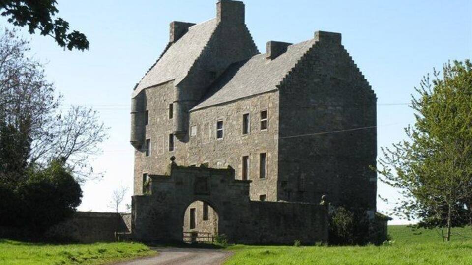 A large stone castle with a prominent archway entrance, surrounded by green fields and trees.