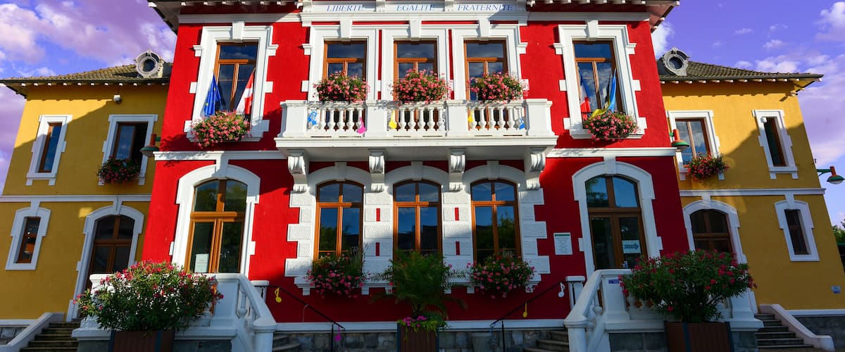 A red and yellow building with a clock tower, balconies with flowers, and a sign reading "Liberté, Égalité, Fraternité".