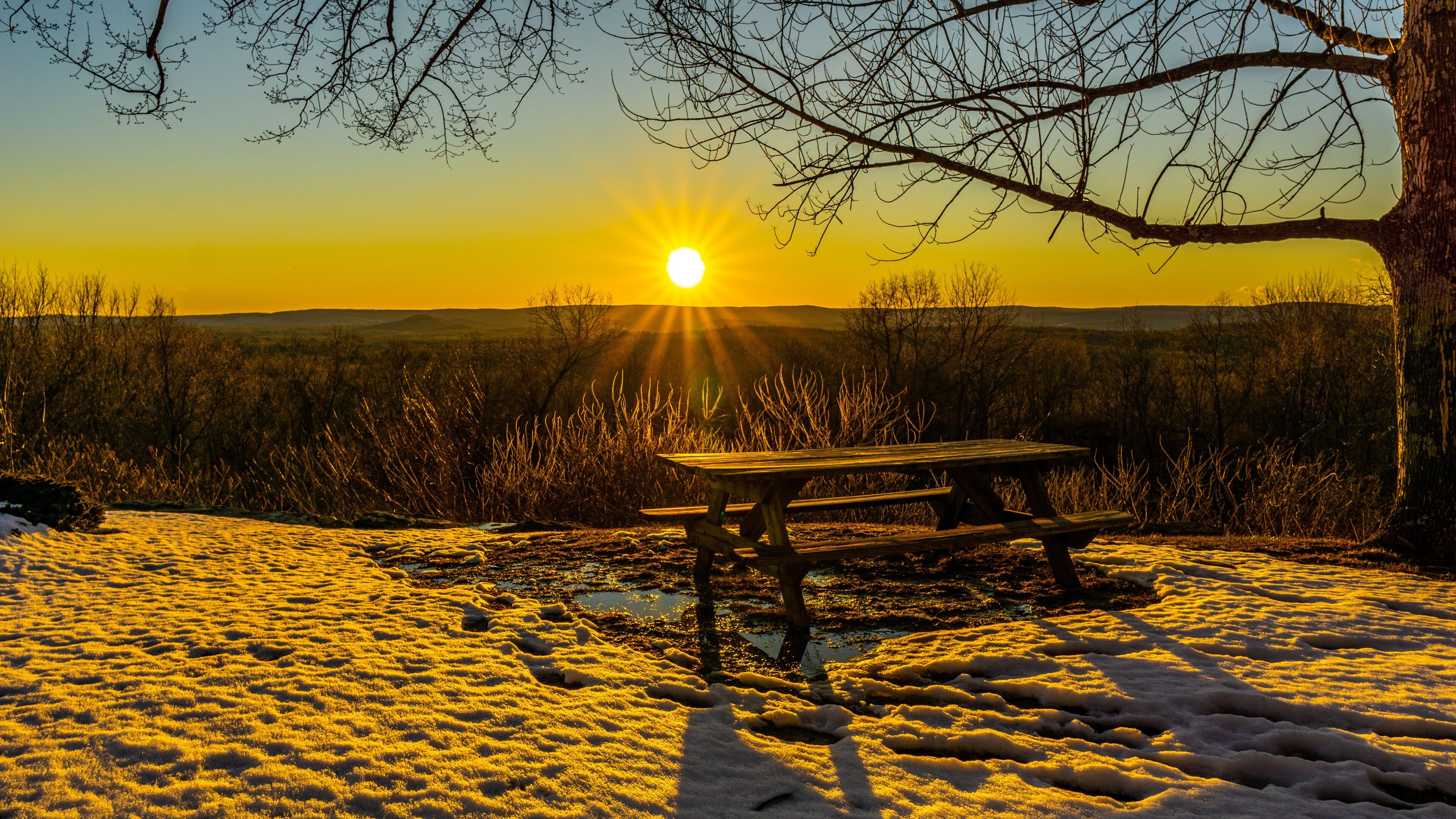Granby Connecticut.  A picnic table on a mountain top rests in snow and mud, while illuminated by a sunset view of another mountain scene.   Beautiful sunset winter scene.