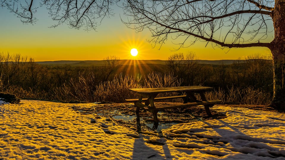 Granby Connecticut. A picnic table on a mountain top rests in snow and mud, while illuminated by a sunset view of another mountain scene. Beautiful sunset winter scene.
