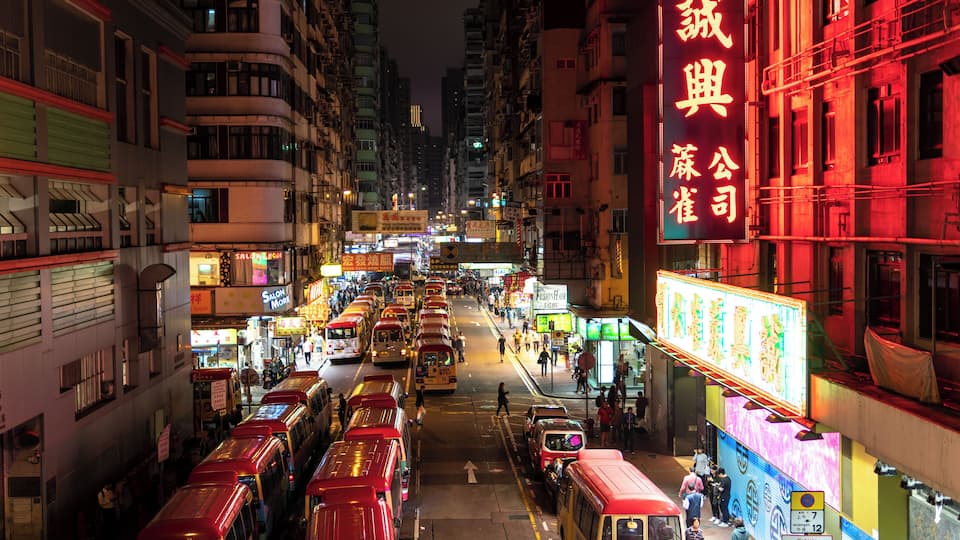 A busy urban street at night with buses, pedestrians, and illuminated signs.