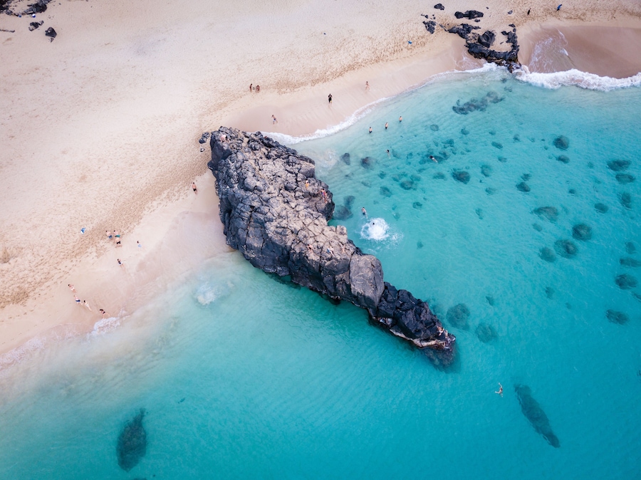 A beach with clear turquoise water, a rocky outcrop, and people swimming and walking on the shore.