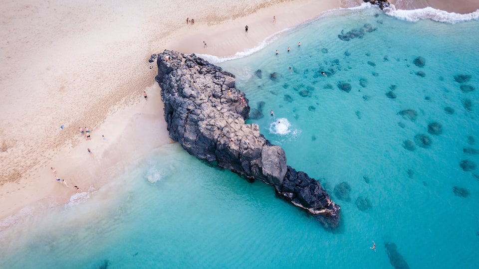 A beach with clear turquoise water, a rocky outcrop, and people swimming and walking on the shore.