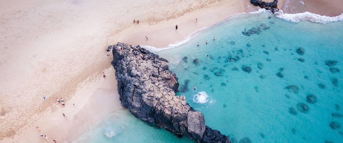 A beach with clear turquoise water, a rocky outcrop, and people swimming and walking on the shore.