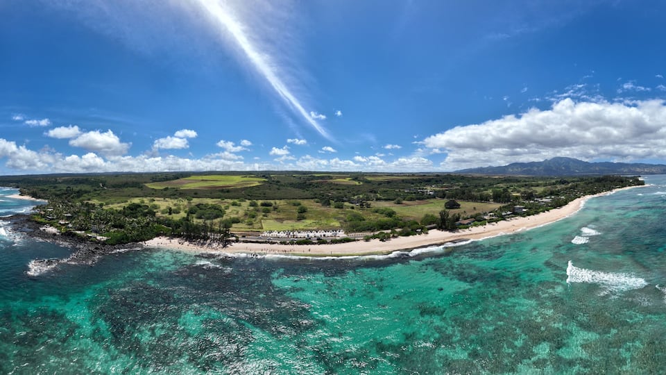 A coastal landscape with a beach, clear turquoise waters, and distant mountains under a blue sky.