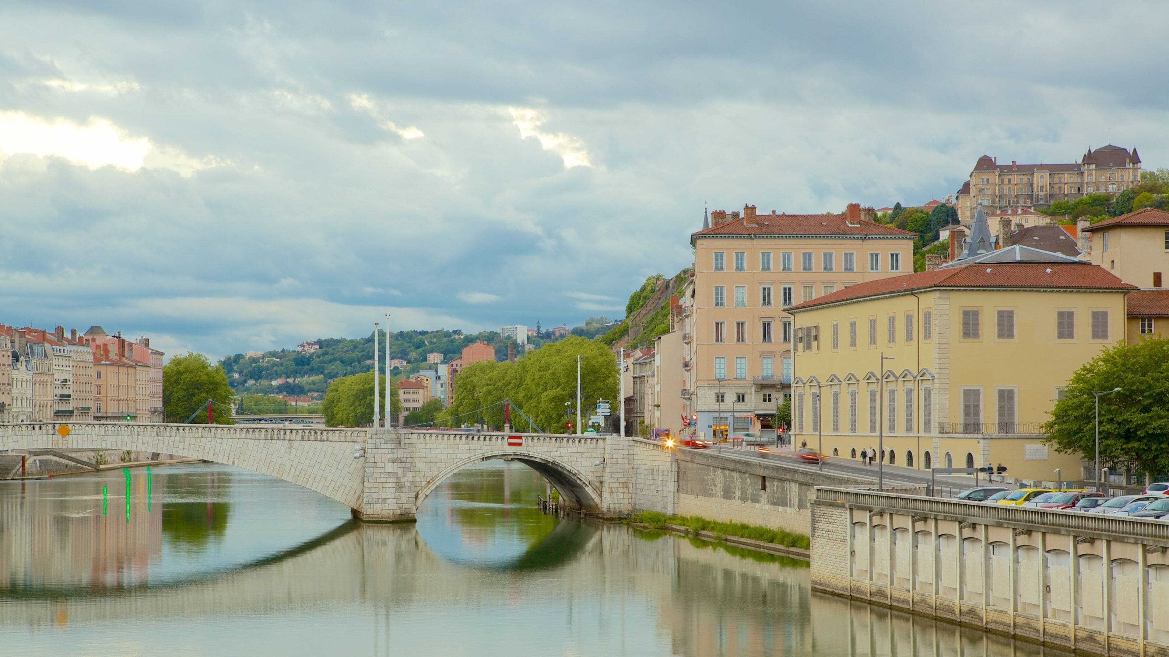A stone bridge over a calm river, surrounded by historic buildings and a hill in the background.