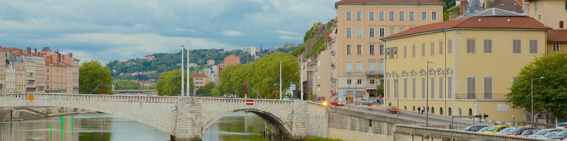 A stone bridge over a calm river, surrounded by historic buildings and a hill in the background.