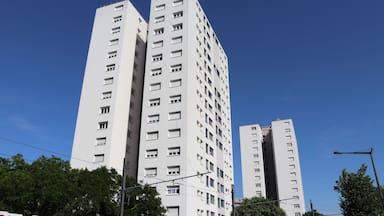 Um edifício alto e branco, com várias janelas e varandas, cercado por árvores sob um céu azul claro.