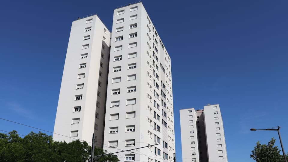 A tall white building with multiple windows and balconies, surrounded by trees and under a clear blue sky.