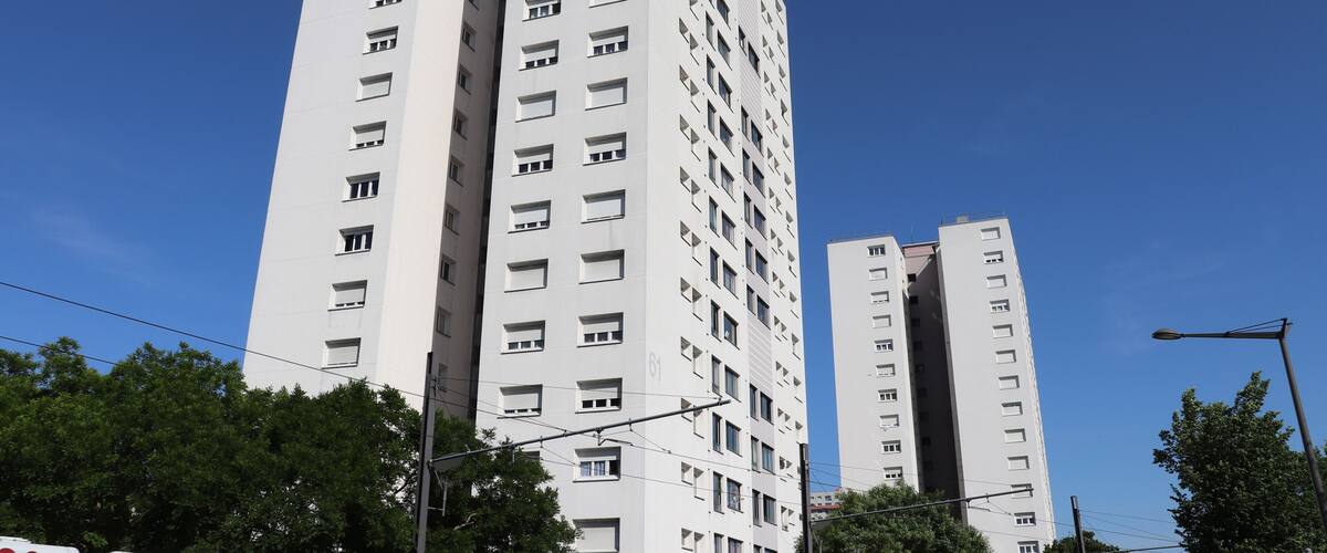 A tall white building with multiple windows and balconies, surrounded by trees and under a clear blue sky.