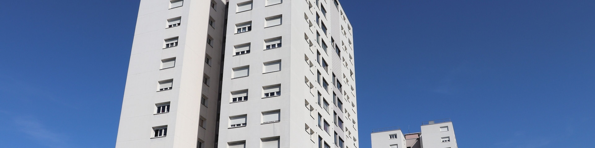 A tall white building with multiple windows and balconies, surrounded by trees and under a clear blue sky.