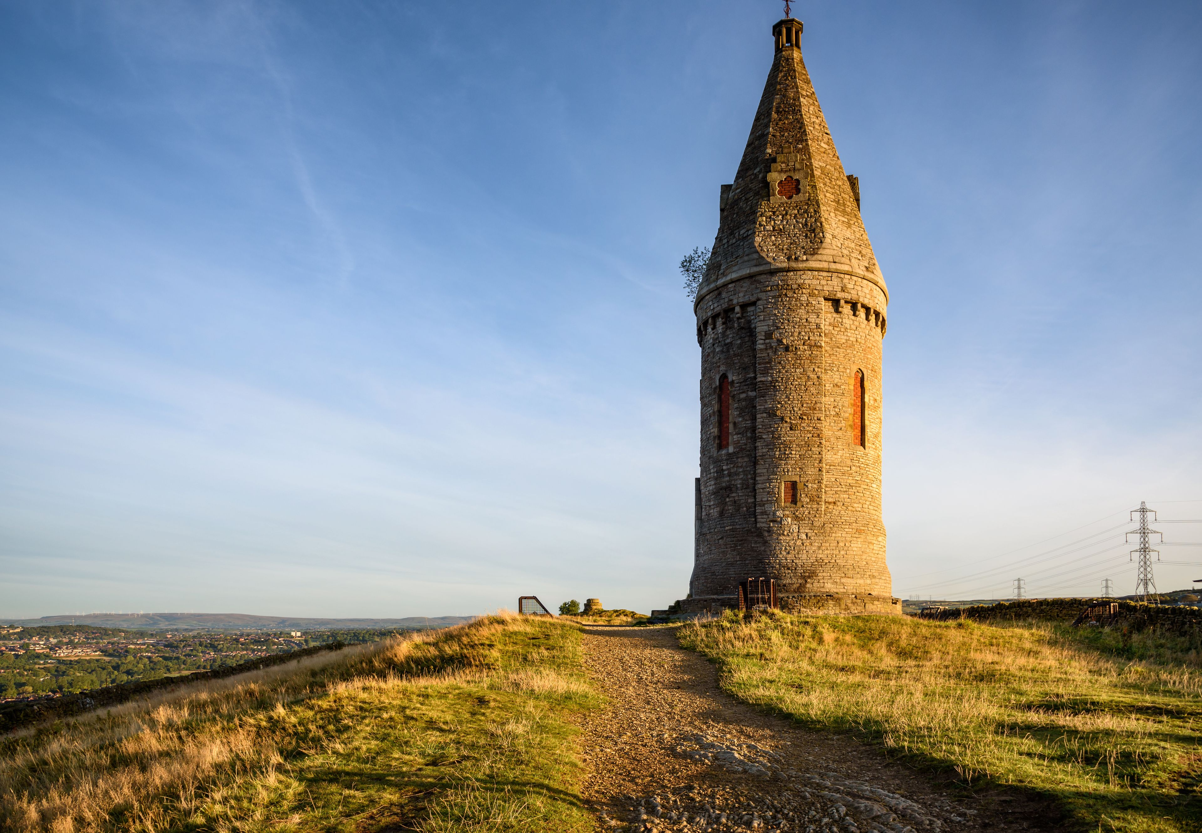 Een hoge stenen toren met een puntig dak, omgeven door grasvelden en een heldere lucht.