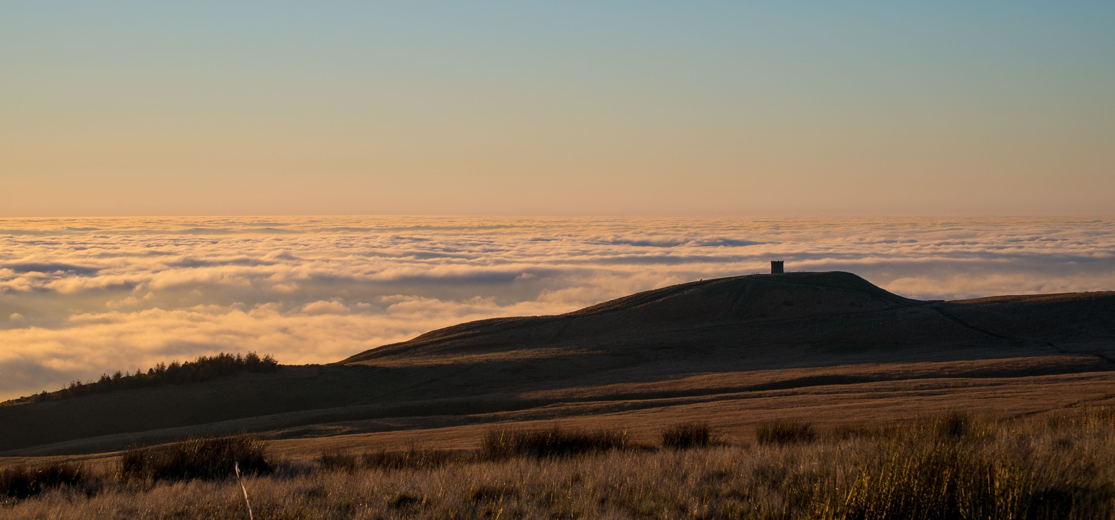 A hill with a tower on top, surrounded by a field and a layer of clouds below.