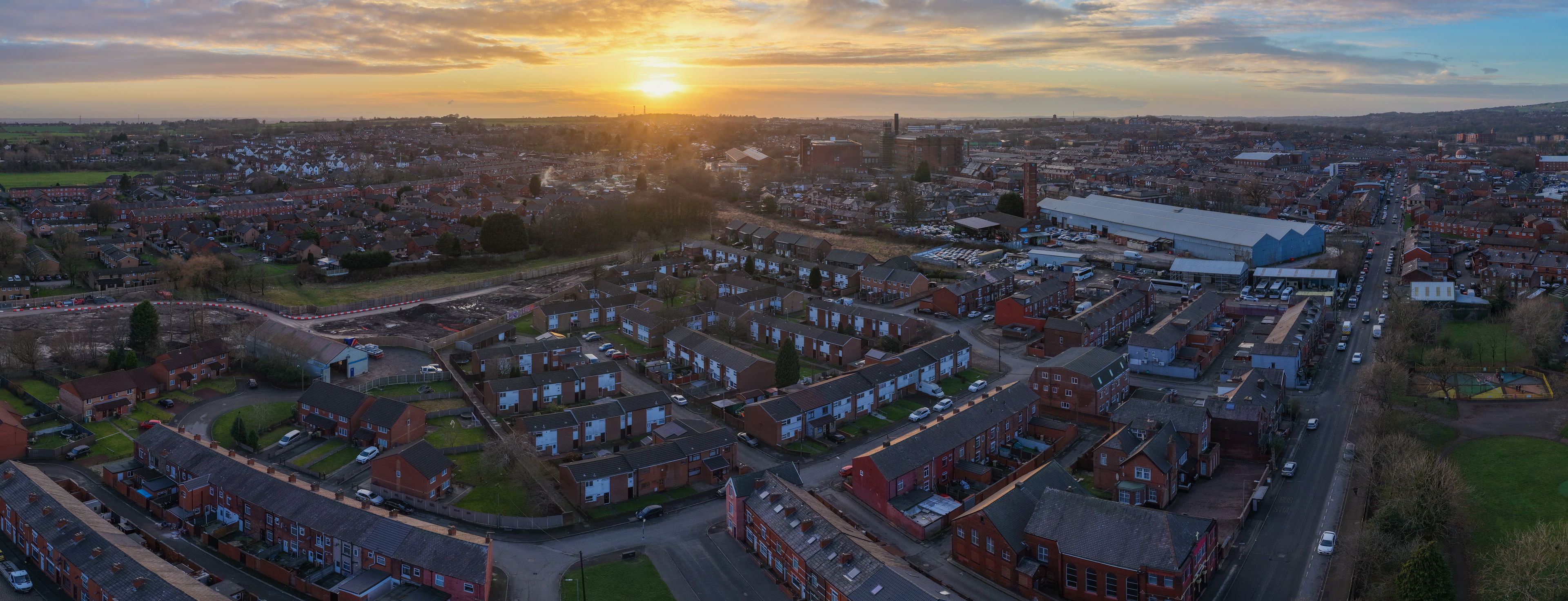 Aerial panorama looking north-west over Bolton, featuring traditional residential streets and industrial heritage against a dramatic Lancashire sunset.