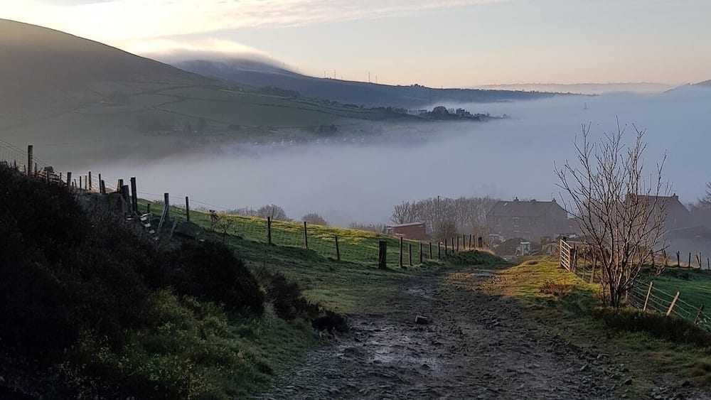 A muddy path leading through a rural landscape with a fence on the left, a tree on the right, and a misty hill in the background.