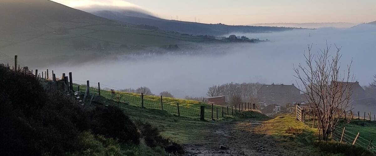 A muddy path leading through a rural landscape with a fence on the left, a tree on the right, and a misty hill in the background.