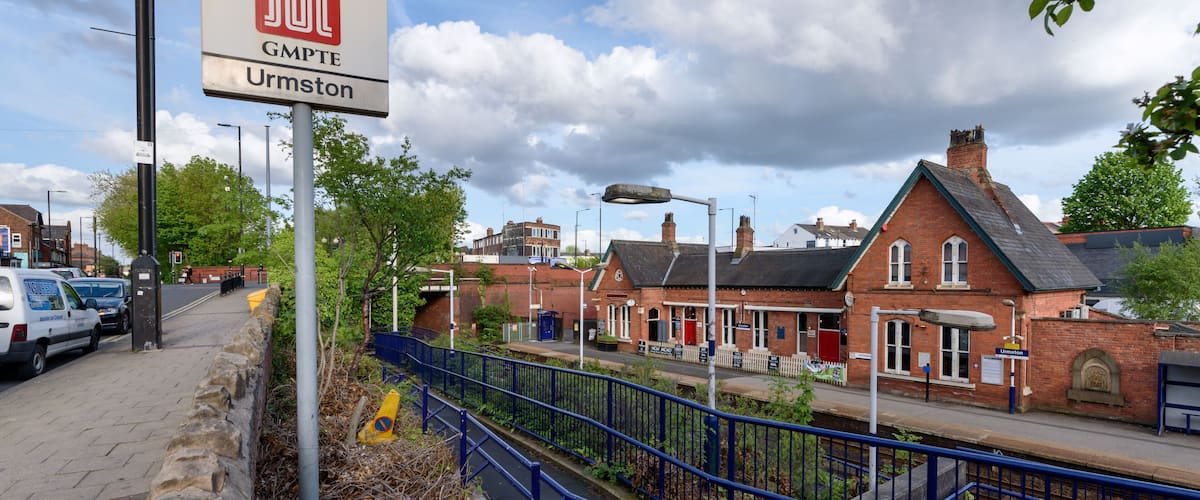 A railway station sign, a stone wall, a blue fence, and a red brick building.