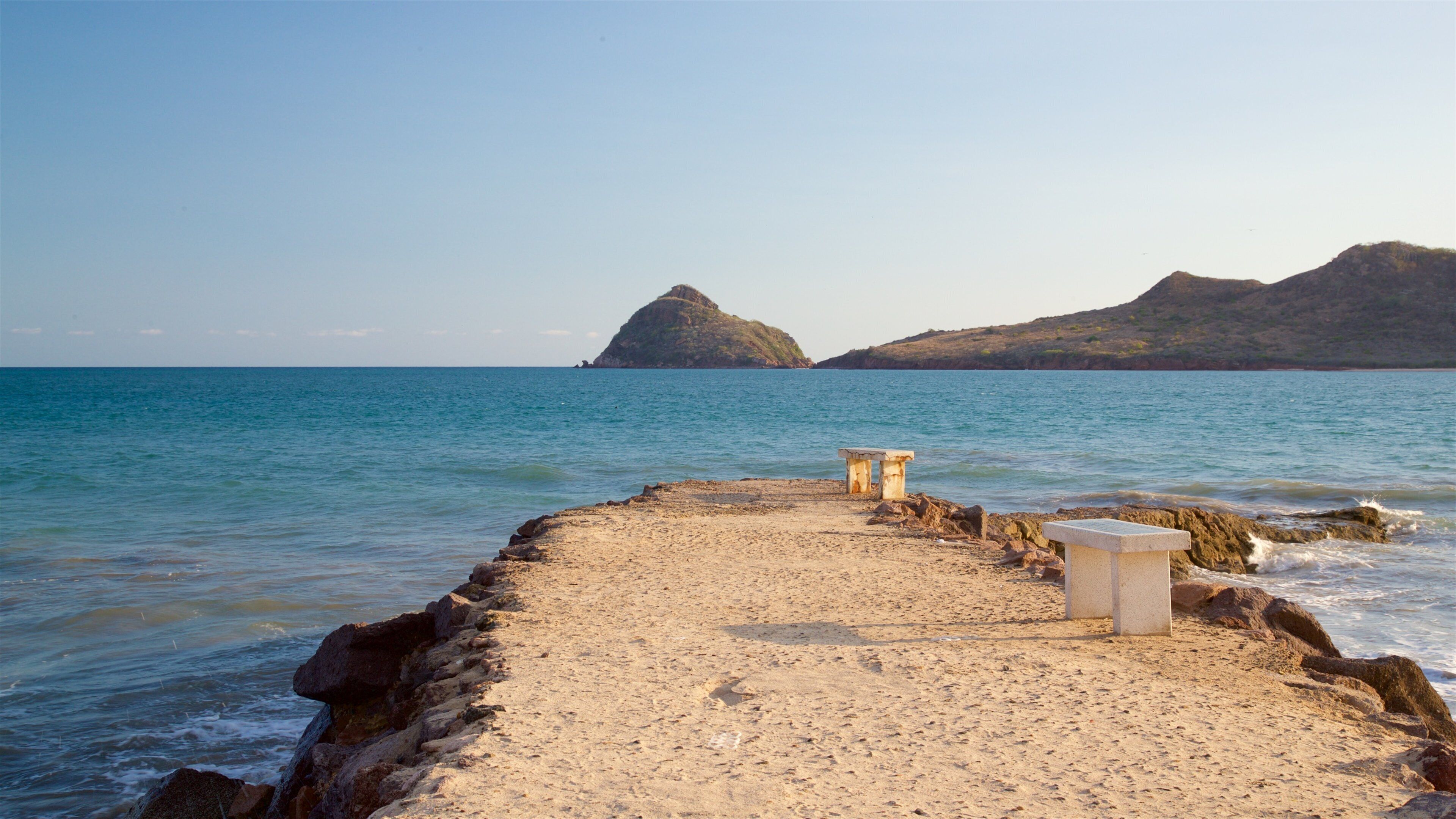 A sandy beach with two stone benches facing the sea, with rocky outcrops and hills in the background.