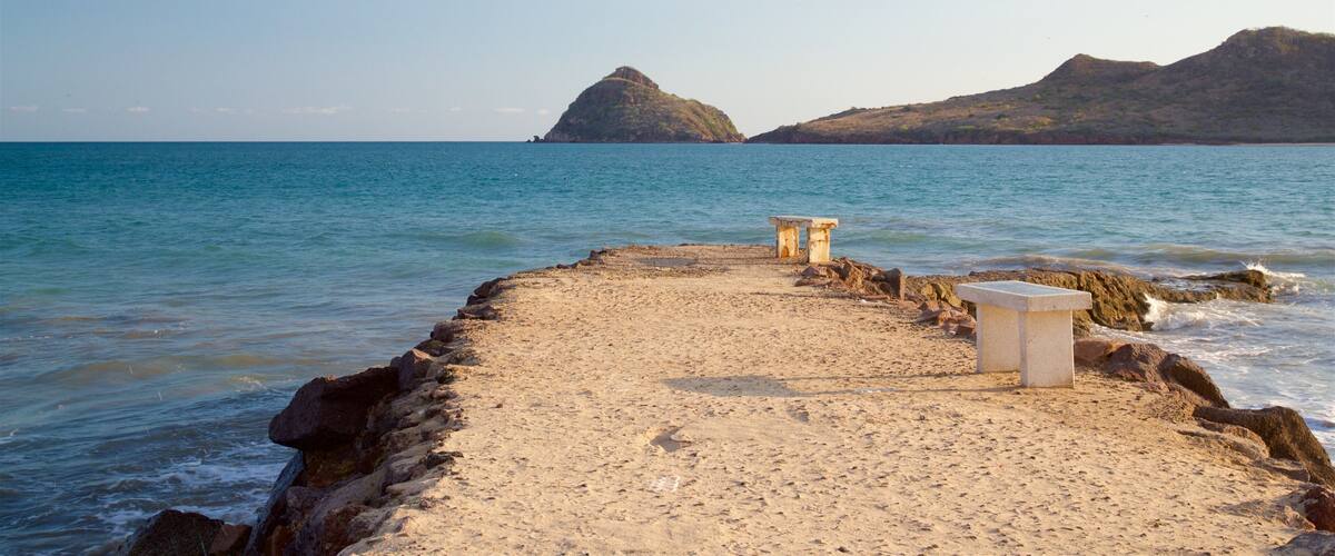 A sandy beach with two stone benches facing the sea, with rocky outcrops and hills in the background.