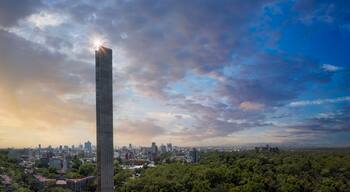 A tall skyscraper with a sunlit top, surrounded by a cityscape and dense green trees.