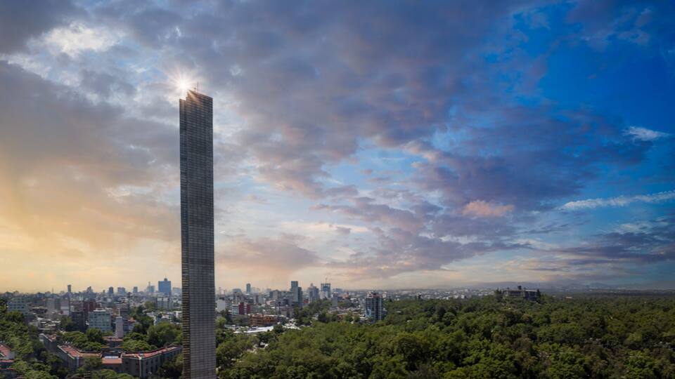 A tall skyscraper with a sunlit top, surrounded by a cityscape and dense green trees.