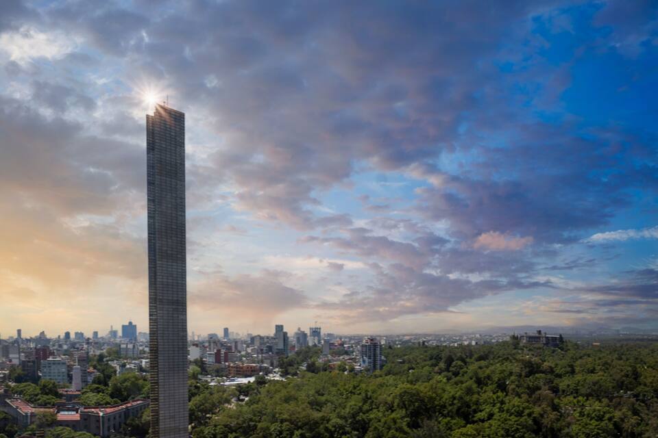 Um arranha-céu imponente com o topo iluminado pelo sol, rodeado por um horizonte urbano e árvores verdes densas.