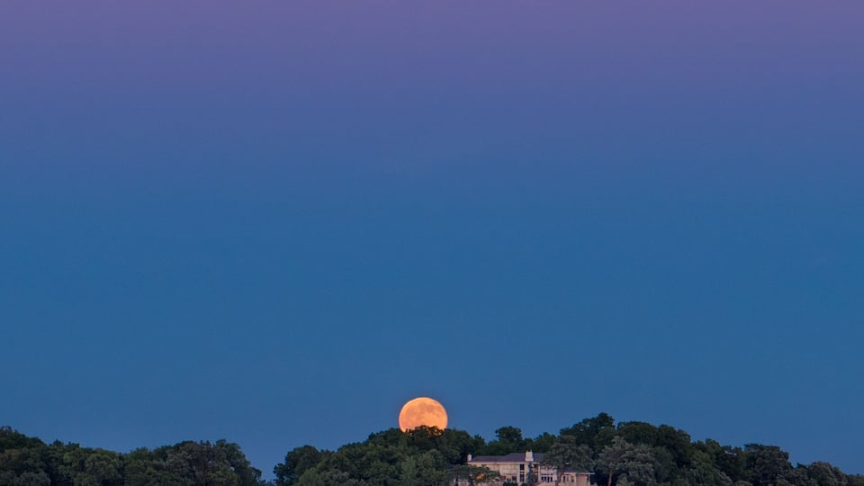 A large moon over a forested hill with buildings at the base.