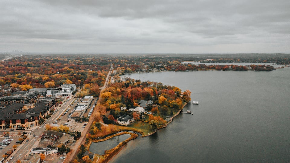 An aerial view of a waterfront area with autumn-colored trees, a marina, and a cityscape in the background.
