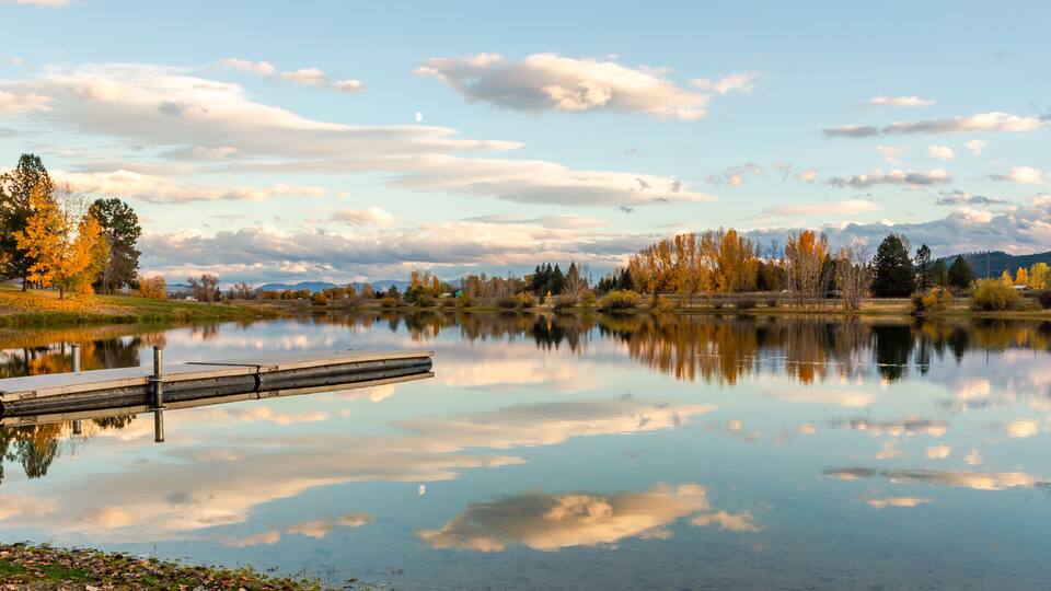 A calm lake with a dock, surrounded by autumn trees and a clear sky.