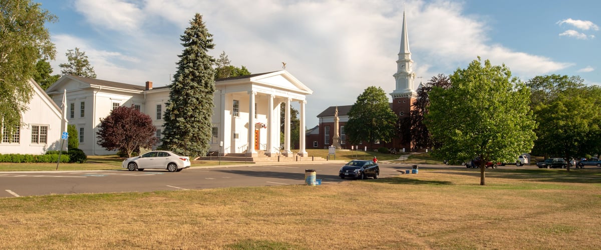 A building with a white facade and columns, a church with a steeple, and a grassy area with trees.
