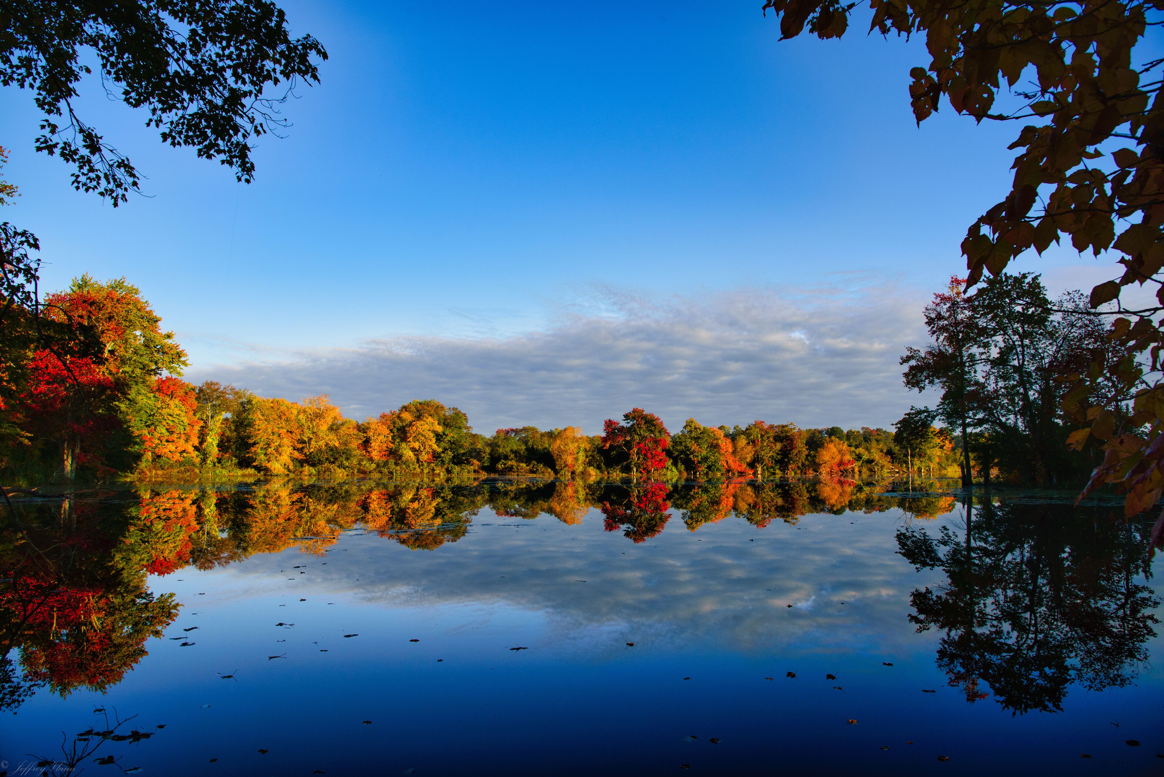 A calm lake reflecting autumn trees with red, orange, and green foliage.