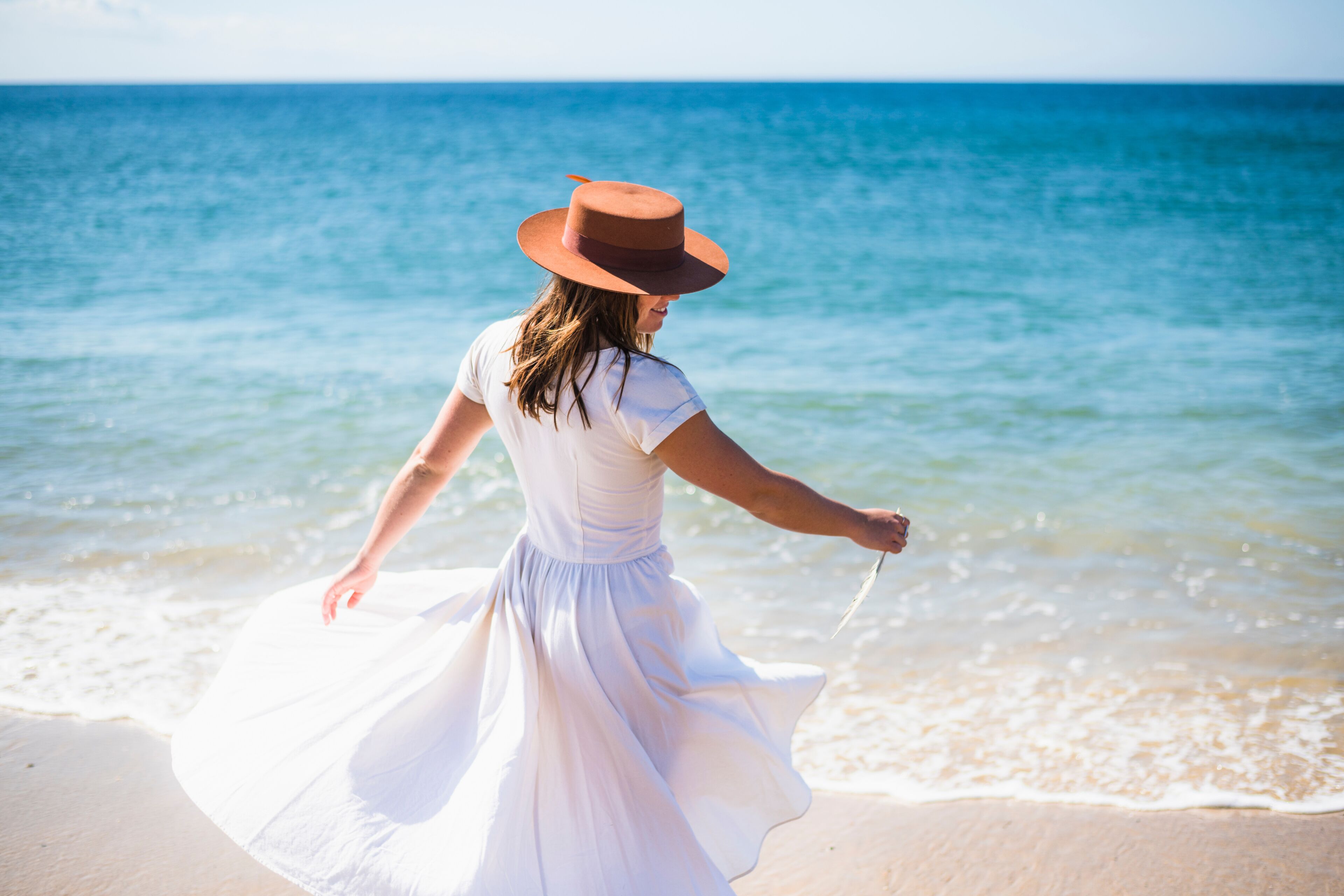 Uma pessoa de vestido branco e chapéu castanho caminhando pela praia, com o oceano ao fundo.