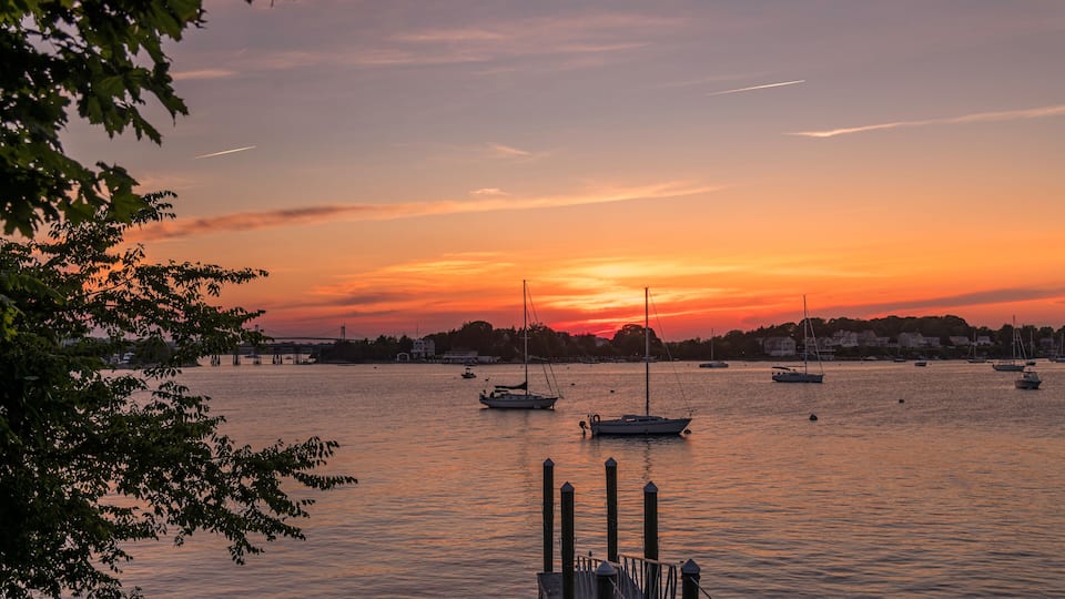 A sunset over a calm harbor with boats and a bridge in the background.