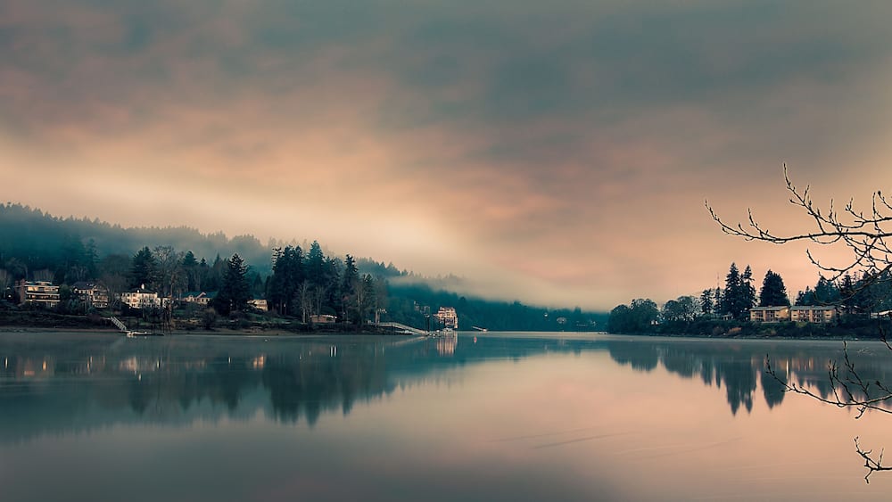 Ein ruhiger See mit Spiegelungen von Häusern und Bäumen, umgeben von bewaldeter Landschaft und bewölktem Himmel.