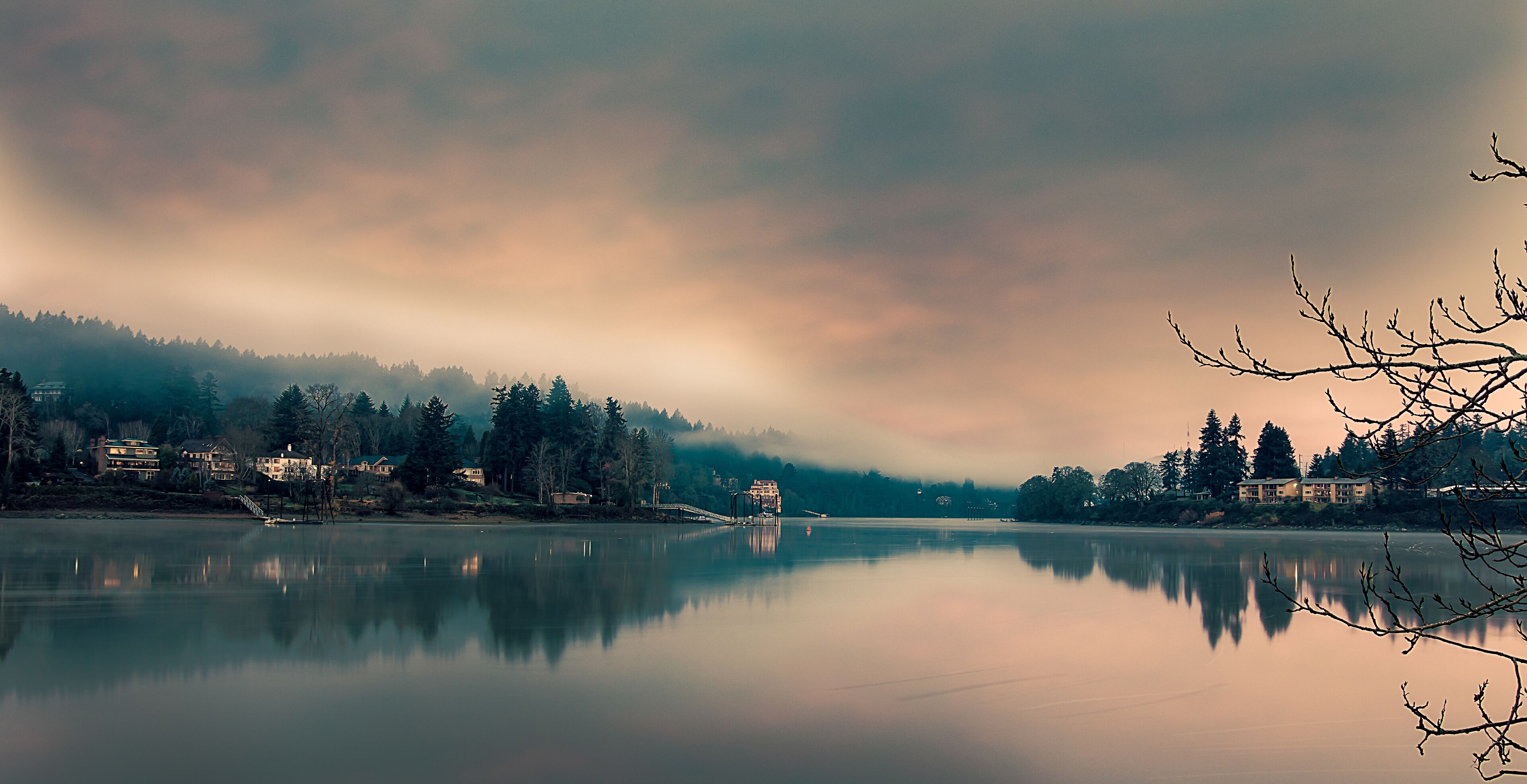 A calm lake with reflections of houses and trees, surrounded by a forested area and a cloudy sky.