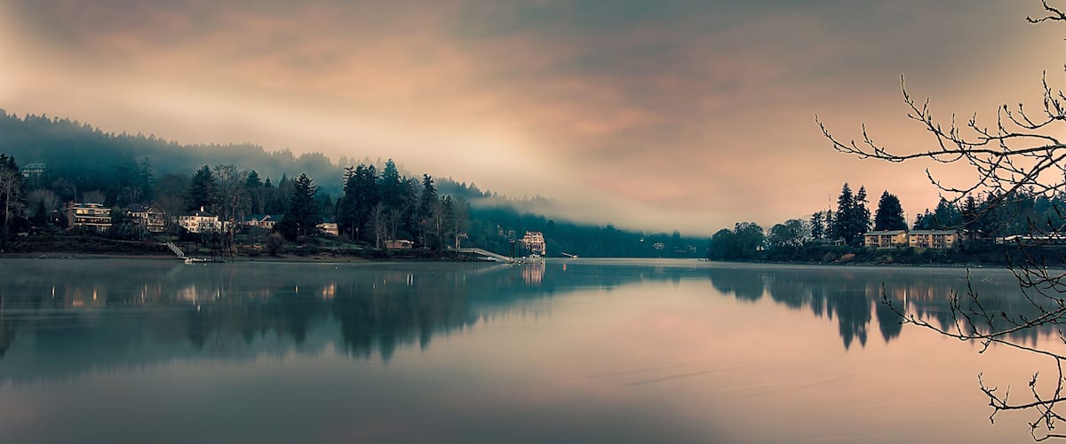 A calm lake with reflections of houses and trees, surrounded by a forested area and a cloudy sky.