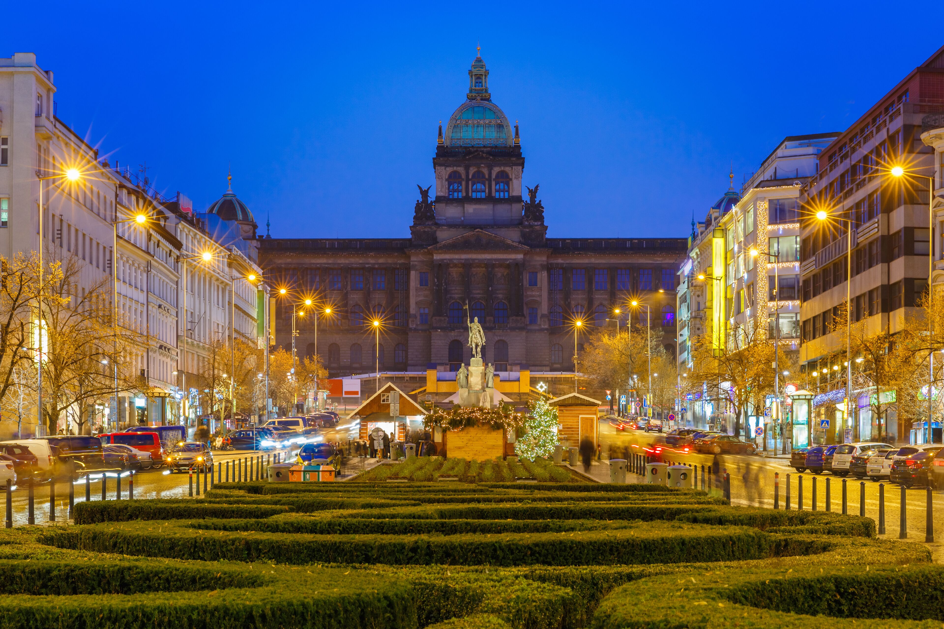 A city square with a central statue, surrounded by manicured hedges and buildings with illuminated facades.