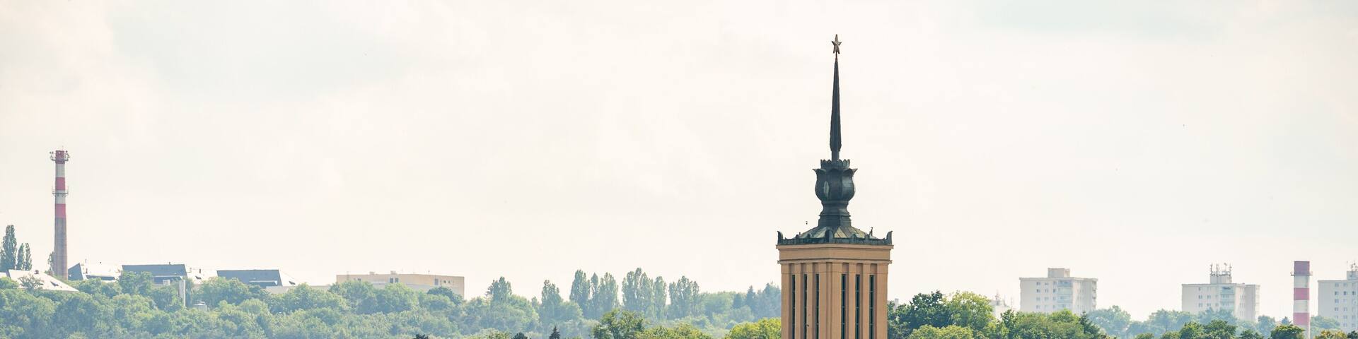 A cityscape with a prominent tower featuring a spire and a statue on top, surrounded by residential buildings and greenery.