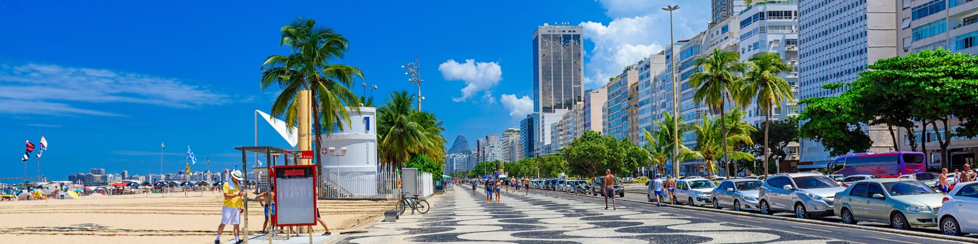 Um calçadão à beira-mar com palmeiras, pavimento estampado e edifícios altos ao fundo.