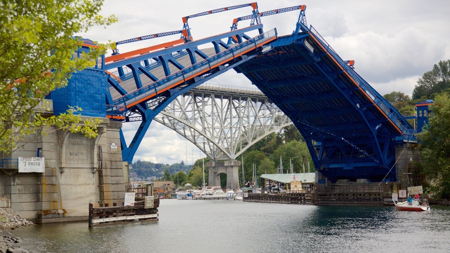 A blue bridge with orange accents over a river, surrounded by trees and boats.