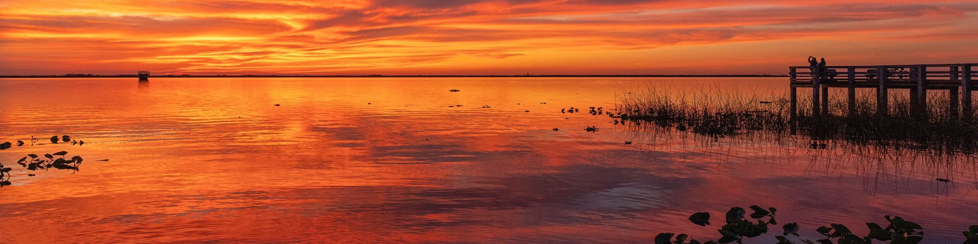 A sunset over a calm body of water with a wooden pier and distant shoreline.