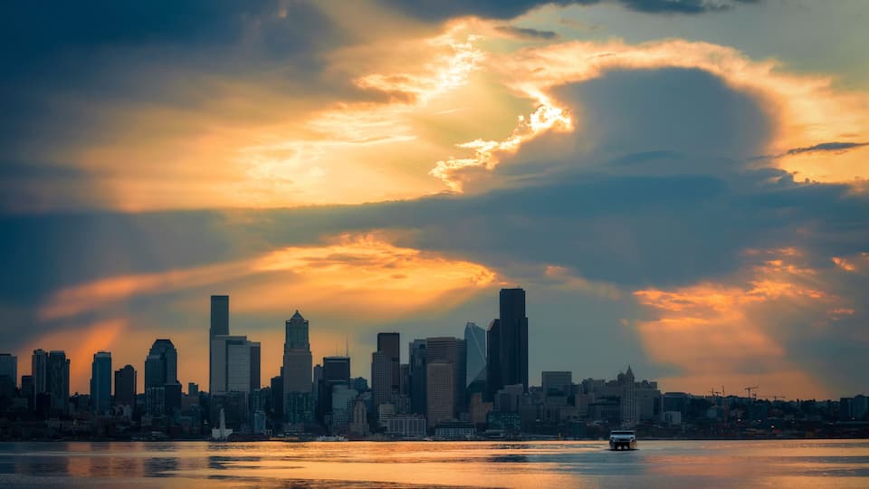 A city skyline at sunset with a river in the foreground and a boat.