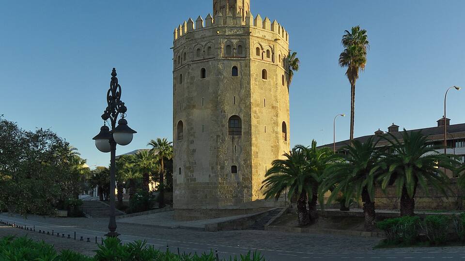 A historic tower with a dome and battlements, surrounded by palm trees and cobblestone pavement.