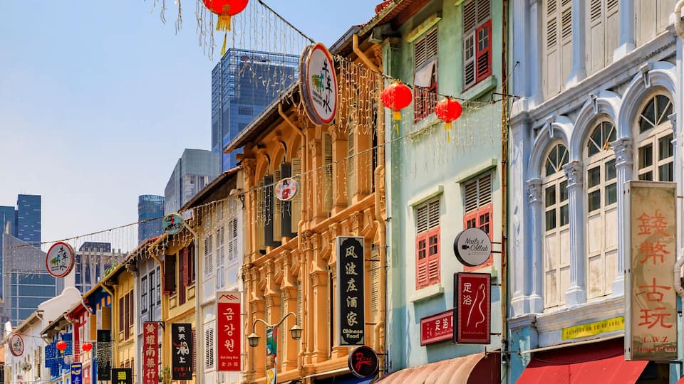 A row of colorful buildings with traditional Chinese signage, a clear blue sky, and modern skyscrapers in the background.