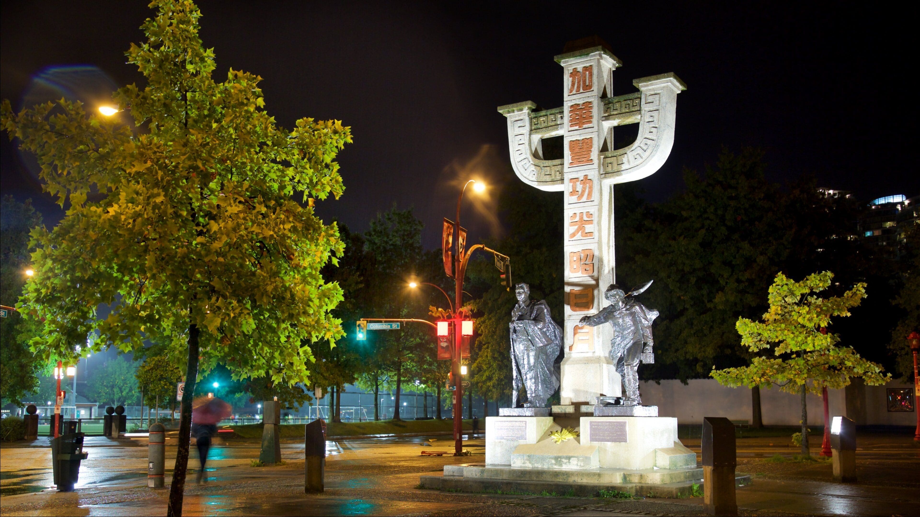 Um monumento em forma de cruz com inscrições, rodeado por árvores e postes de luz à noite.