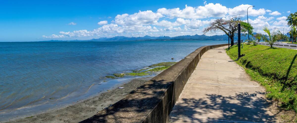 A coastal walkway with a clear view of the sea, mountains in the distance, and a concrete barrier separating the path from the water.