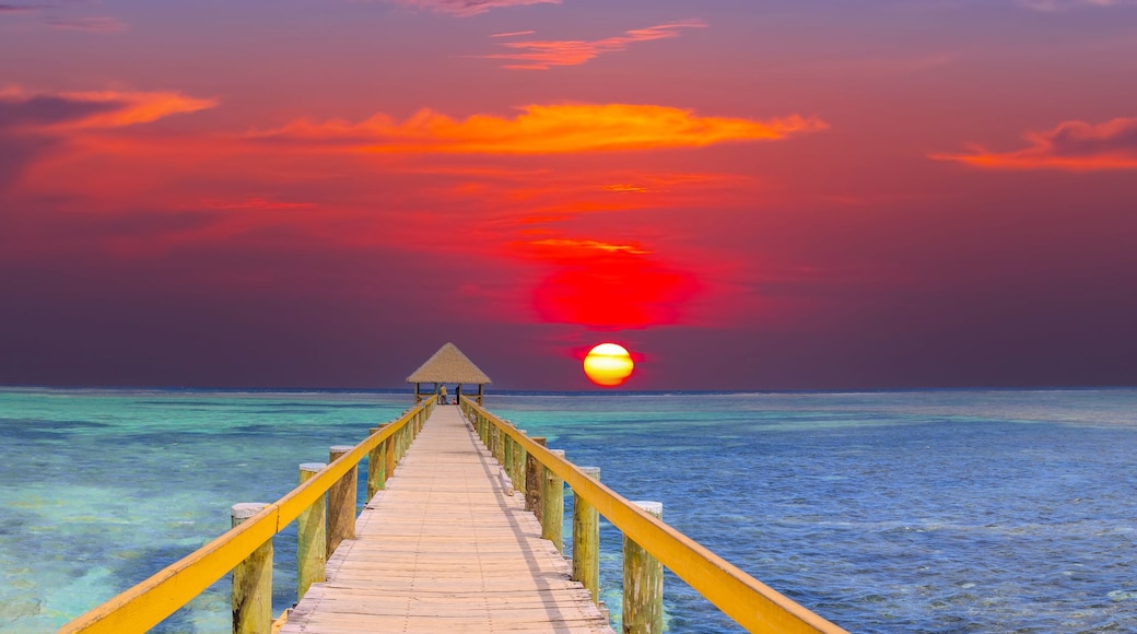 A wooden pier leading to a thatched-roof structure on a tropical beach with clear turquoise waters and a sunset.