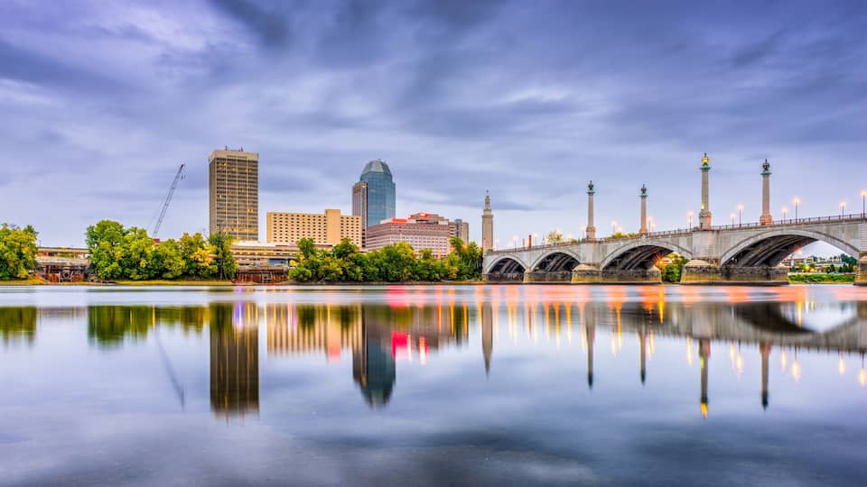A city skyline with a river reflecting buildings, a bridge with multiple arches, and a tower.