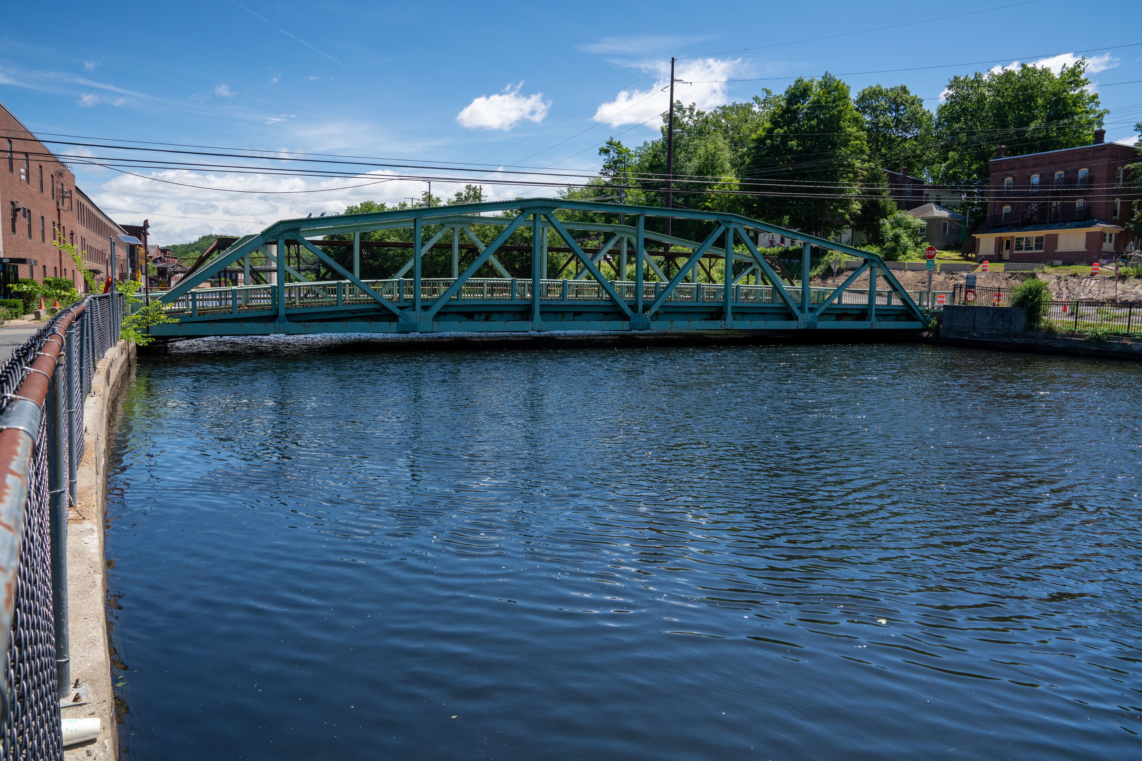 A turquoise metal bridge over a calm river, surrounded by buildings and trees.