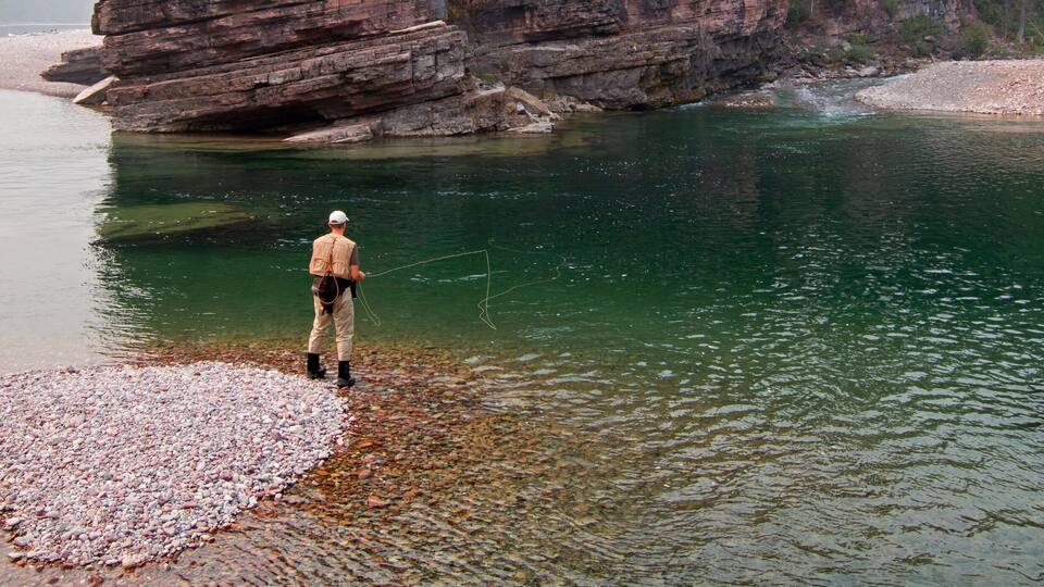 A person fishing by a clear river with rocky banks and a large rock formation in the background.